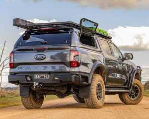 a loaded touring ute (HiLux or Ranger with canopy, bull bar, roof rack) in a regional or bush setting